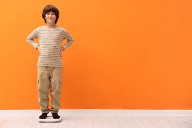 Photo of Little boy standing on scales near orange wall indoors. Space for text