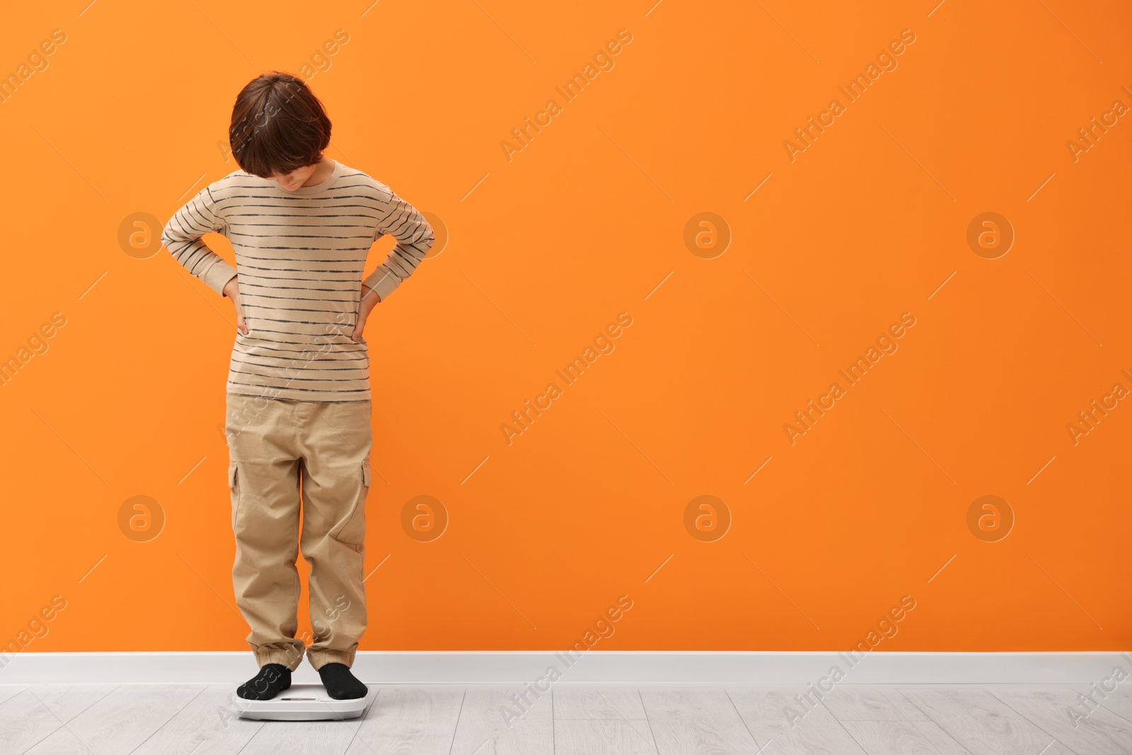 Photo of Little boy standing on scales near orange wall indoors. Space for text