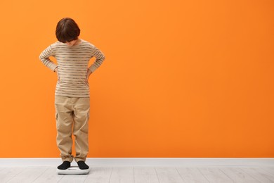 Photo of Little boy standing on scales near orange wall indoors. Space for text