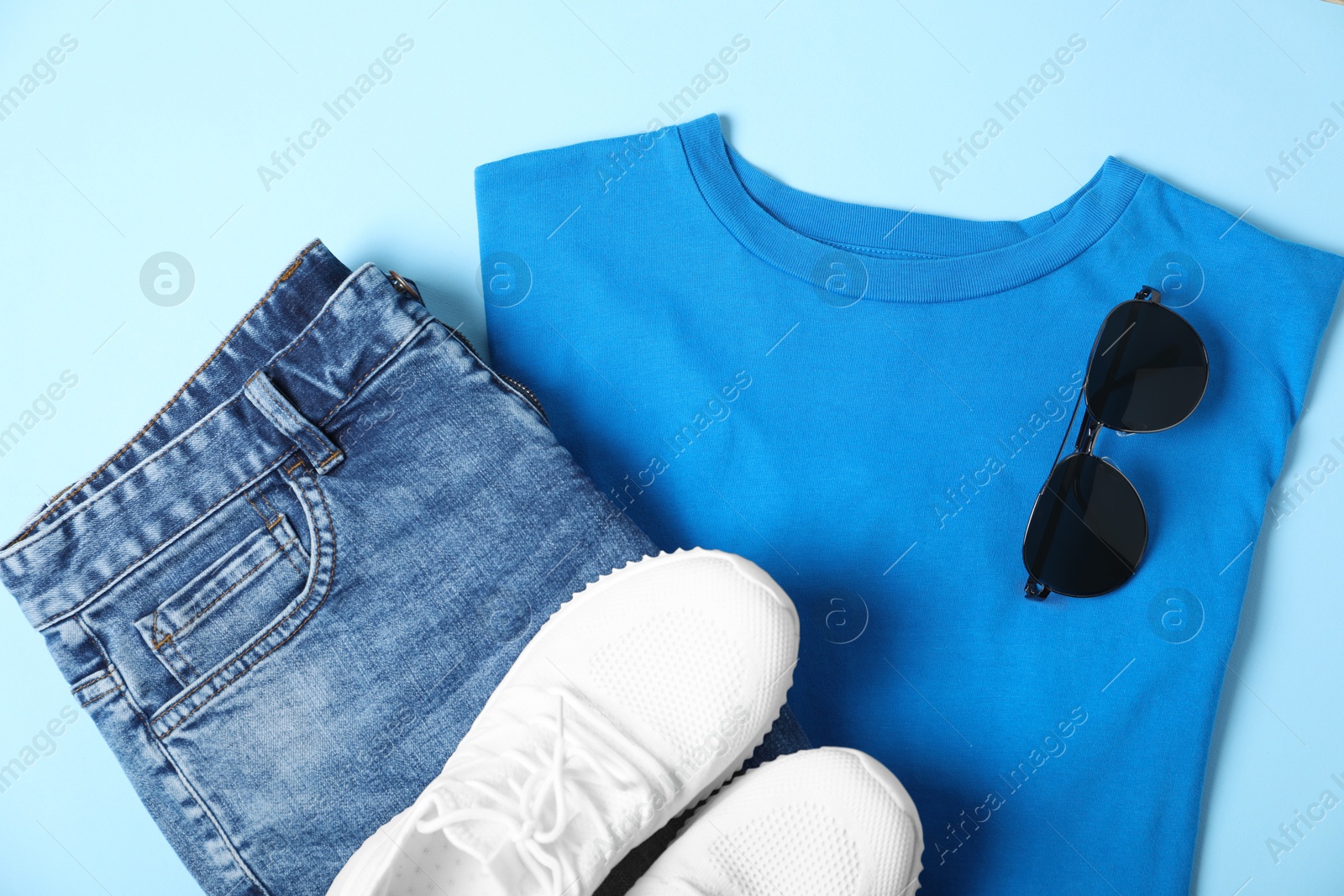 Photo of T-shirt, jeans, sunglasses and sneakers on light blue background, flat lay. Mockup for design