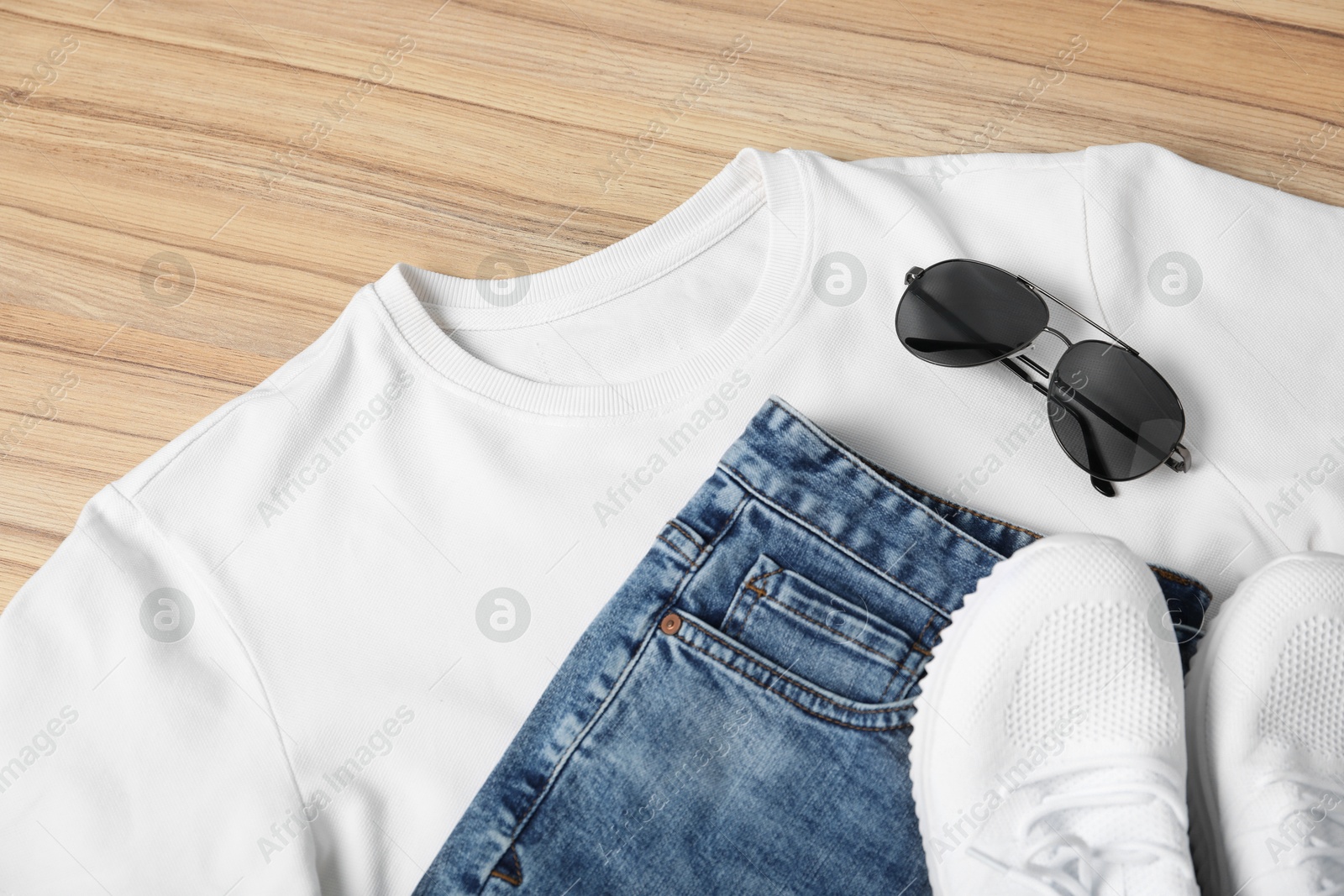 Photo of White t-shirt, jeans, sunglasses and sneakers on wooden table, flat lay. Mockup for design