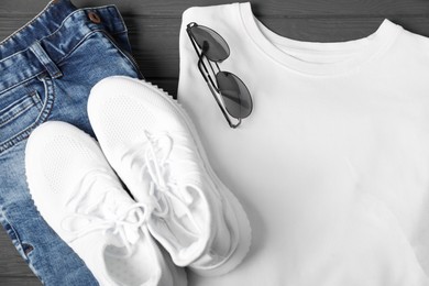 Photo of White t-shirt, jeans, sunglasses and sneakers on grey wooden table, flat lay. Mockup for design