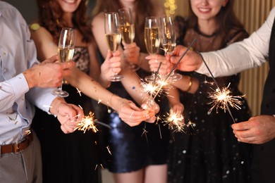 Photo of Friends celebrating New Year with sparklers and wine glasses indoors, closeup