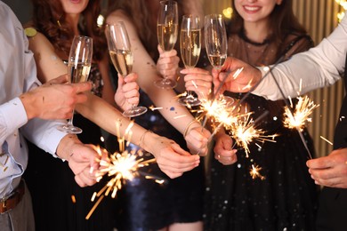 Photo of Friends celebrating New Year with sparklers and wine glasses indoors, closeup