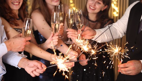 Photo of Friends celebrating New Year with sparklers and wine glasses indoors, selective focus