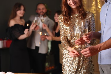 Photo of Friends celebrating New Year with sparklers and wine glasses indoors, closeup