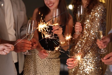 Photo of Friends celebrating New Year with sparklers and wine glasses indoors, closeup