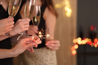 Photo of Women celebrating New Year with sparklers and wine glasses indoors, closeup. Space for text