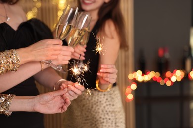 Photo of Women celebrating New Year with sparklers and wine glasses indoors, closeup
