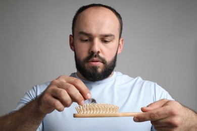 Photo of Baldness problem. Worried man with brush and clump of lost hair on grey background