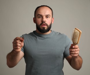 Photo of Baldness problem. Worried man with brush and clump of lost hair on grey background
