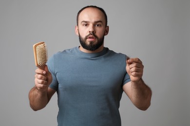 Photo of Baldness problem. Worried man with brush and clump of lost hair on grey background
