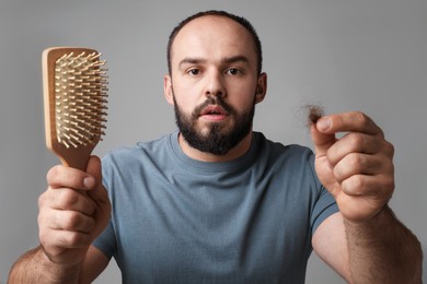 Photo of Baldness problem. Worried man with brush and clump of lost hair on grey background