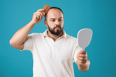 Photo of Baldness problem. Man brushing his hair with comb and looking at mirror on light blue background