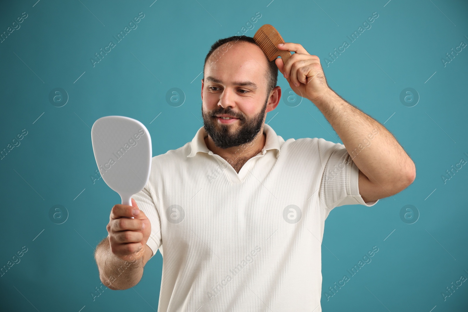 Photo of Baldness problem. Smiling man brushing his hair with comb and looking at mirror on light blue background