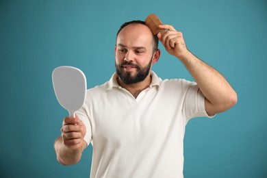 Photo of Baldness problem. Smiling man brushing his hair with comb and looking at mirror on light blue background
