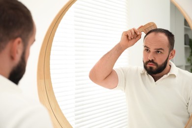 Photo of Baldness problem. Man brushing his hair with comb near mirror at home