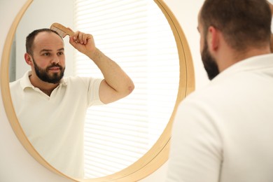 Photo of Baldness problem. Man brushing his hair with comb near mirror at home