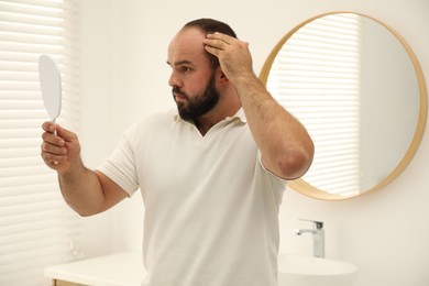 Photo of Baldness problem. Man with receding hairline looking at mirror indoors