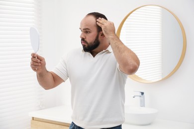Photo of Baldness problem. Man with receding hairline looking at mirror indoors