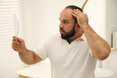 Photo of Baldness problem. Man with receding hairline looking at mirror indoors