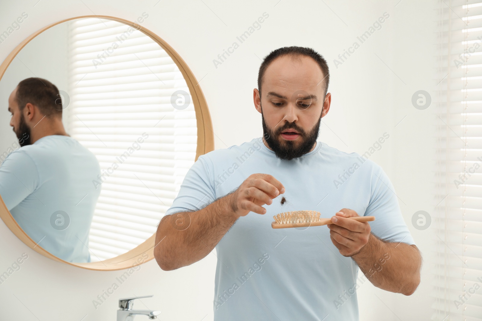 Photo of Baldness problem. Worried man taking his lost hair from brush near mirror in bathroom