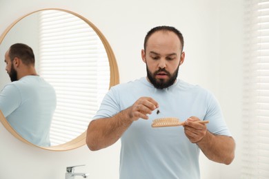 Photo of Baldness problem. Worried man taking his lost hair from brush near mirror in bathroom