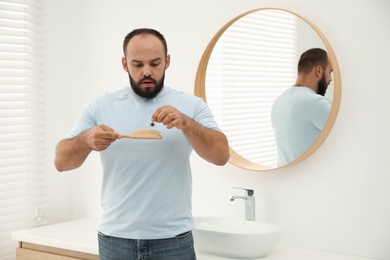 Photo of Baldness problem. Worried man taking his lost hair from brush near mirror in bathroom
