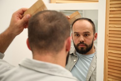 Photo of Baldness problem. Man brushing his hair with comb near mirror at home