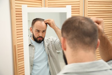 Photo of Baldness problem. Man with receding hairline near mirror at home