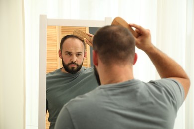 Photo of Baldness problem. Man brushing his hair with comb near mirror at home