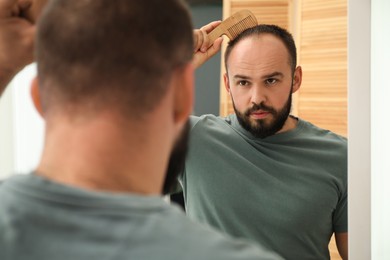 Photo of Baldness problem. Man brushing his hair with comb near mirror at home