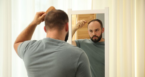 Photo of Baldness problem. Man brushing his hair with comb near mirror at home