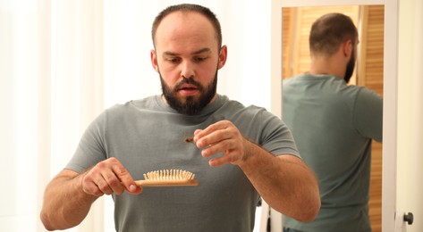 Photo of Baldness problem. Worried man taking his lost hair from brush at home