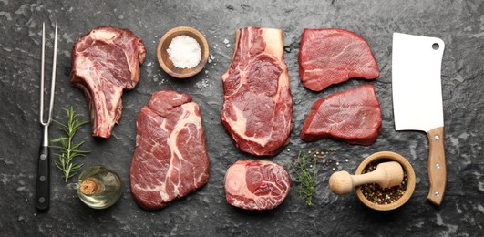 Photo of Pieces of raw beef meat, spices, carving fork and cleaver on black marble table, flat lay