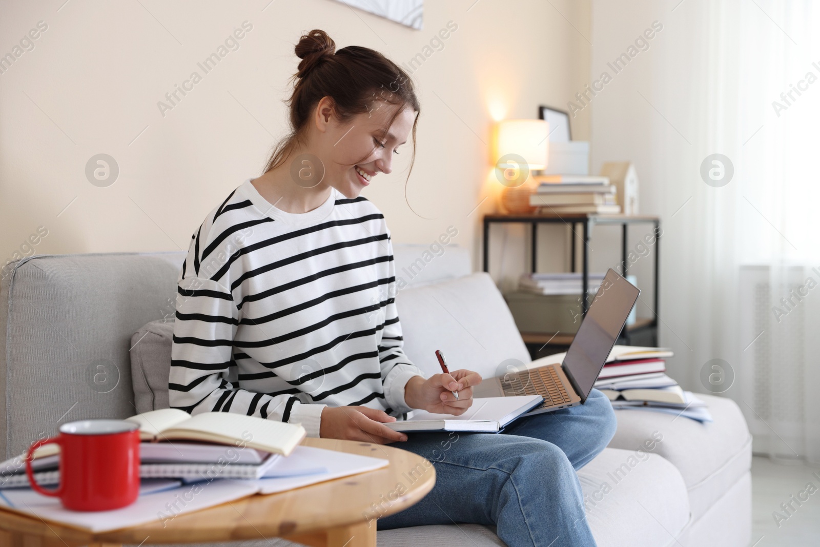Smiling student with laptop, notebook and pen preparing for exam on sofa at home Photo of Smiling student with laptop, notebook and pen preparing for exam on sofa at home