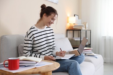 Photo of Smiling student with laptop, notebook and pen preparing for exam on sofa at home