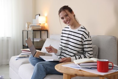 Photo of Smiling student with laptop, notebook and pen preparing for exam on sofa at home