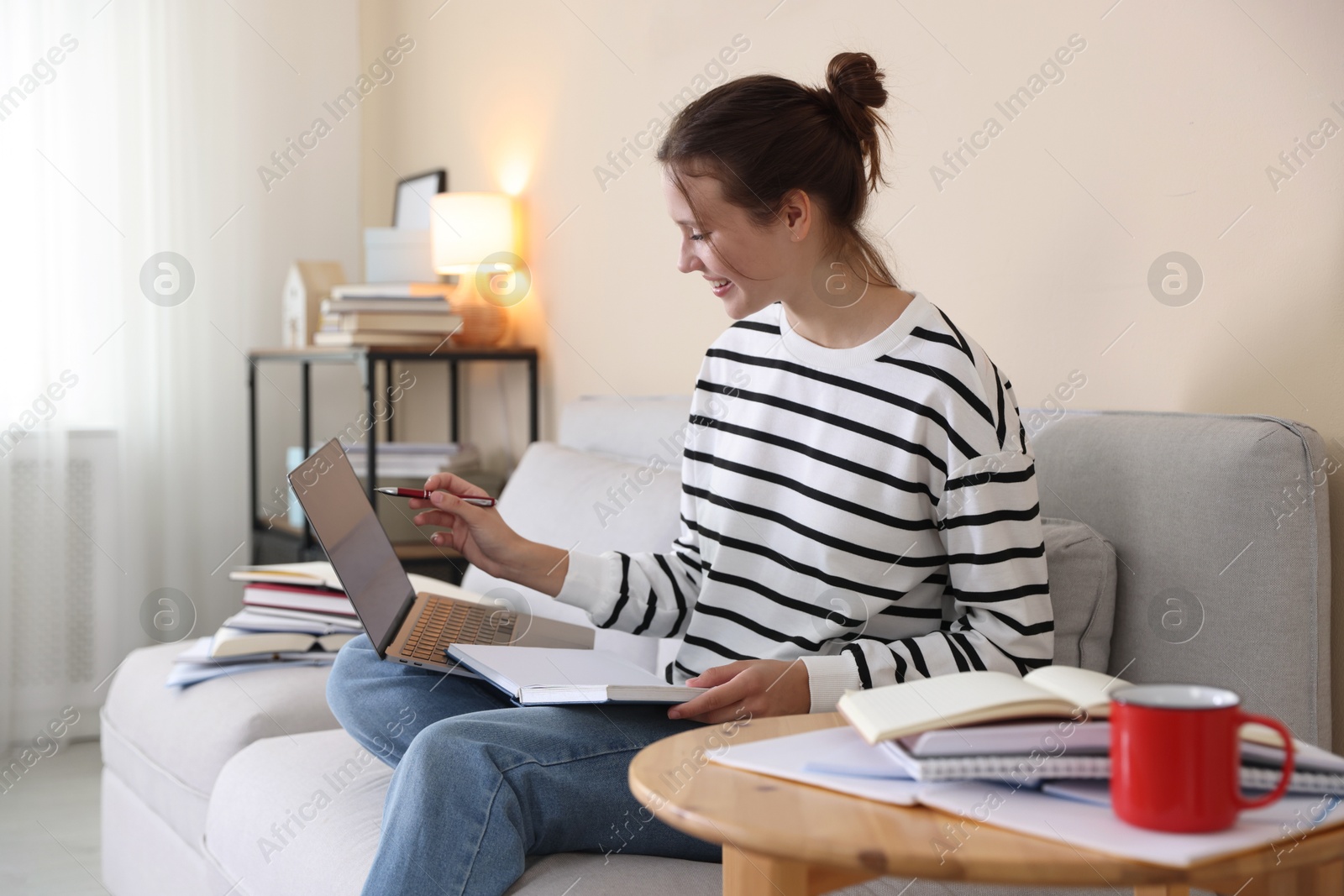 Smiling student with laptop, notebook and pen preparing for exam on sofa at home Photo of Smiling student with laptop, notebook and pen preparing for exam on sofa at home