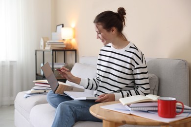 Photo of Smiling student with laptop, notebook and pen preparing for exam on sofa at home
