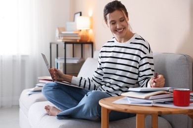 Photo of Smiling student with laptop, notebook and pen preparing for exam on sofa at home