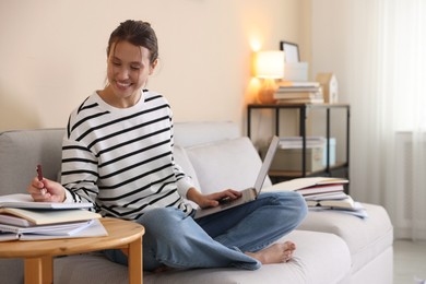Photo of Smiling student with laptop, notebook and pen preparing for exam on sofa at home