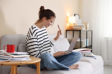 Photo of Smiling student with laptop and pen preparing for exam on sofa at home