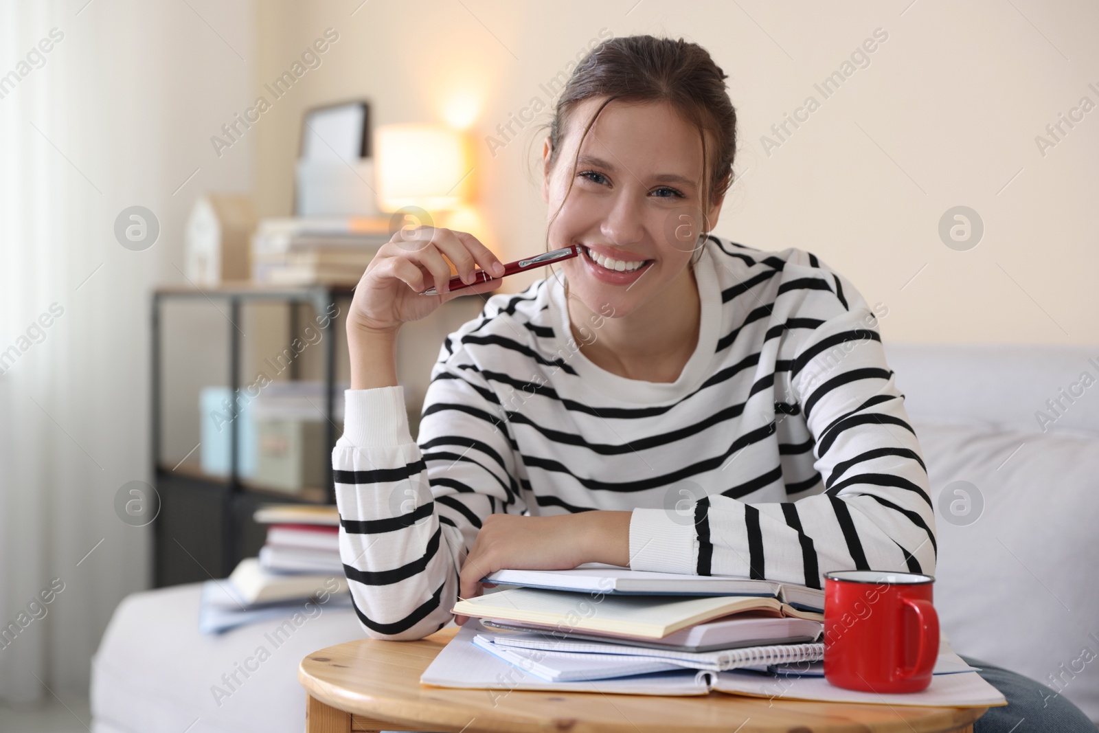 Smiling student at table with books and mug preparing for exam on sofa indoors Photo of Smiling student at table with books and mug preparing for exam on sofa indoors