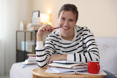 Photo of Smiling student at table with books and mug preparing for exam on sofa indoors