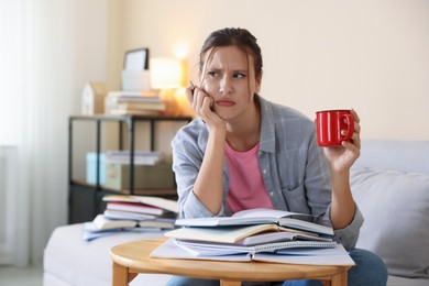 Photo of Worried student at table with books and mug preparing for exam on sofa indoors. Space for text