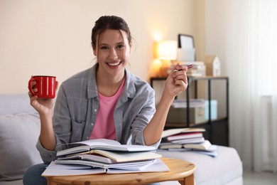 Photo of Smiling student with pen and mug at table with books preparing for exam on sofa indoors