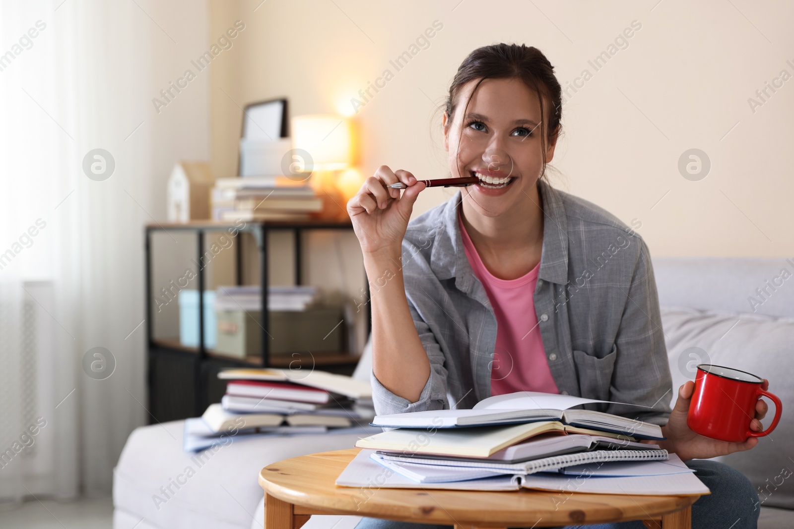 Smiling student with pen and mug at table with books preparing for exam on sofa indoors. Space for text Photo of Smiling student with pen and mug at table with books preparing for exam on sofa indoors. Space for text