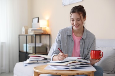 Photo of Smiling student at table with books and mug preparing for exam on sofa indoors. Space for text