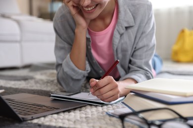 Photo of Smiling student with laptop, notebook and pen preparing for exam on floor indoors, closeup